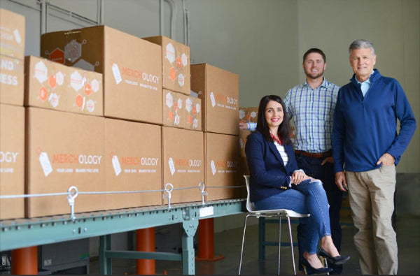 Standing in front of a conveyor belt of boxes, the executive board of Merchology celebrate being a disability-owned business