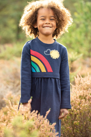 Little girl standing in a field of dried grass wearing the Leia Loopback applique dress in Indigo Rainbow by Frugi Organics. Dress has half of a rainbow on one side and an applique sun with thread rays on the other, and the skirt is complete with POCKETS! 