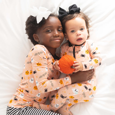 An older girl laying on a bed snuggling a baby - older girl is wearing the Macaron + Me Halloween print pajamas and the baby is wearing the footies in the same print, holding a pumpkin. Both girls are wearing bows. 