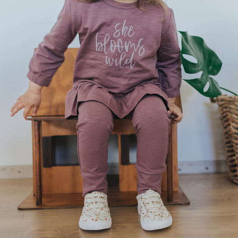 Baby wearing the She Blooms Wild peplum tee and the matching wildberry leggings from Buck & Baa - sitting on a chair with a view of just the outfit and shoes. 