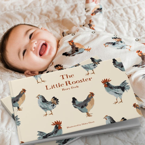 MilkBarn Kids Chicken print footed romper on a little baby laying on the new chicken print organic cotton crib sheet with the new The Little Rooster Book, arriving early spring 2021. 