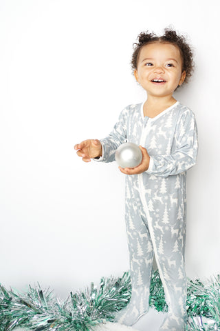 Toddler girl standing in a lifestyle photoshoot for Emerson and Friends wearing the White Christmas holiday print bamboo footie pajama, smiling big in front of a winter wonderland setup. 
