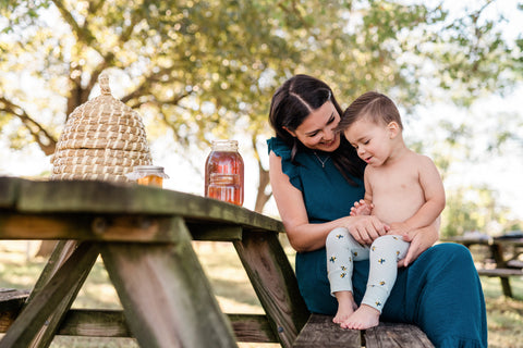 Milkbarn Kids lifestyle photo of little toddler boy wearing new Spring 2021 bumblebee print bamboo leggings while sitting at picnic table on mom's lap with jar of honey