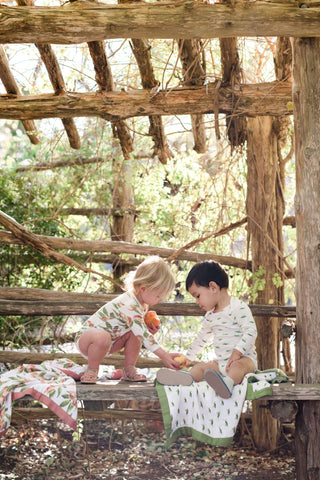 Milkbarn Kids Lifestyle Photo of two toddlers wearing new organic cotton prints, Grasshopper and Peaches, while sharing a peach on the Grasshopper Big Lovey Blanket in the romper and long sleeve one piece bodysuit. 