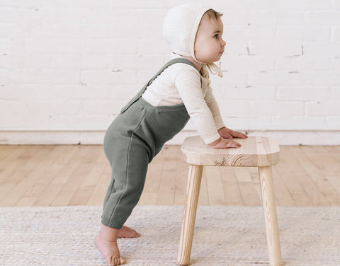 Baby wearing Quincy Mae Knit overalls in basil with a pixie bonnet in natural while leaning on a wooden stool. 