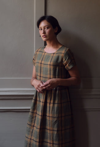 a model wearing a green and brown plaid linen dress stands in front of an old, georgian, wooden paneled wall