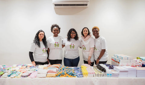 5 women arm in arm wearing colourful beginning charity tshirts, in front of items being donated to premature babies