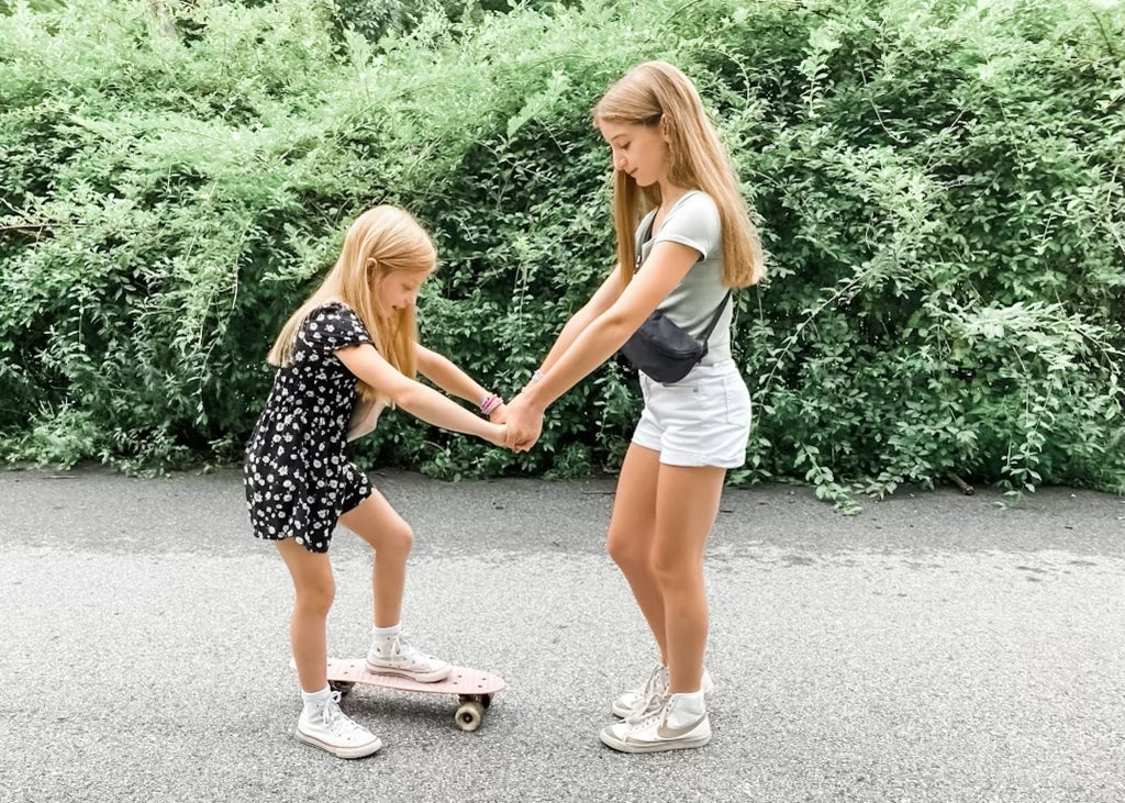 Sisters holding hands learning to skateboard wearing Kibou Mini fanny packs