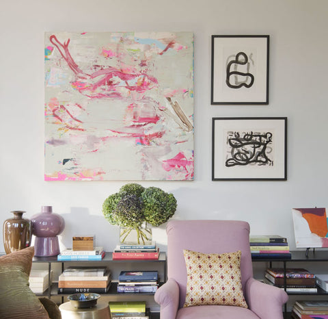Wall of a living room, with a large abstract piece of art hanging on the wall, complemented by two smaller pieces. A bookshelf against the wall obscured by a light purple arm chair