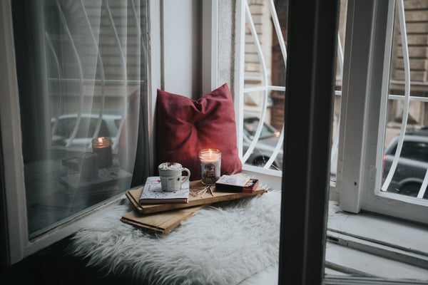 An open window sill, painted white, with a furry white pillow sitting next to the open window. A red pillow sits next to the window, in front of it a mug filled with hot chocolate, a lit candle, some matches and a matchbook, a book, and some window plants. Photo by Alisa Anton on Unsplash.