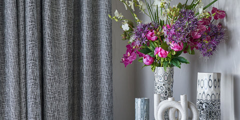 Photo of a room with a gray curtain on the left side, and on the right are several neutral colored vases, one with a bouquet of flowers in a range of greens and pinks.