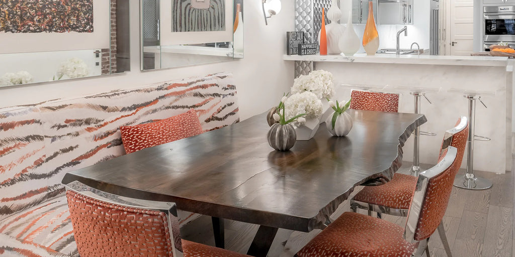 A organically shaped wooden dining table is bordered by a number of orange upholstered chairs and a white bench with stripes of orange and gray. In the background, a white counter with stools, and a number of decorative accessories sit on top, with a kitchen in the background.