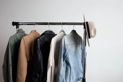 A clothes rack stands against a wall, with 5 different jackets hanging on it and a hat dangling off the right side
