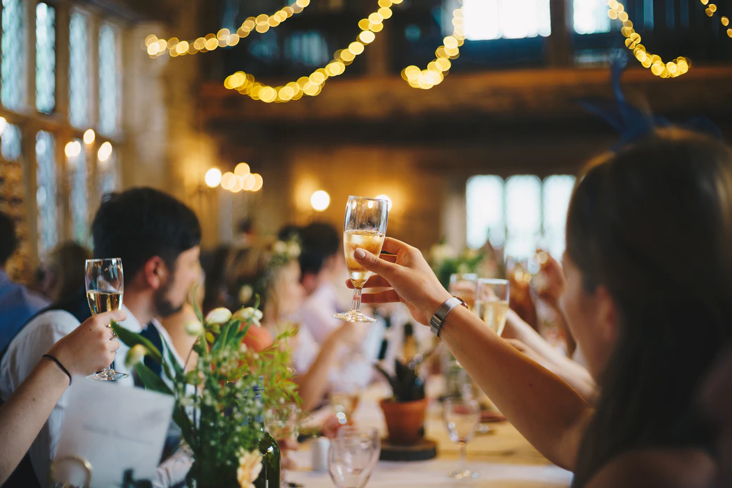 In focus in front is a person with long hair holding a full champagne flute up for a toast, with another hand coming in from the left edge of the photo doing the same. Other blurred people in the background are also raising their hands and all looking towards the horizon of the picture. The people are in formal clothes sitting at a table with glasses and flowers.