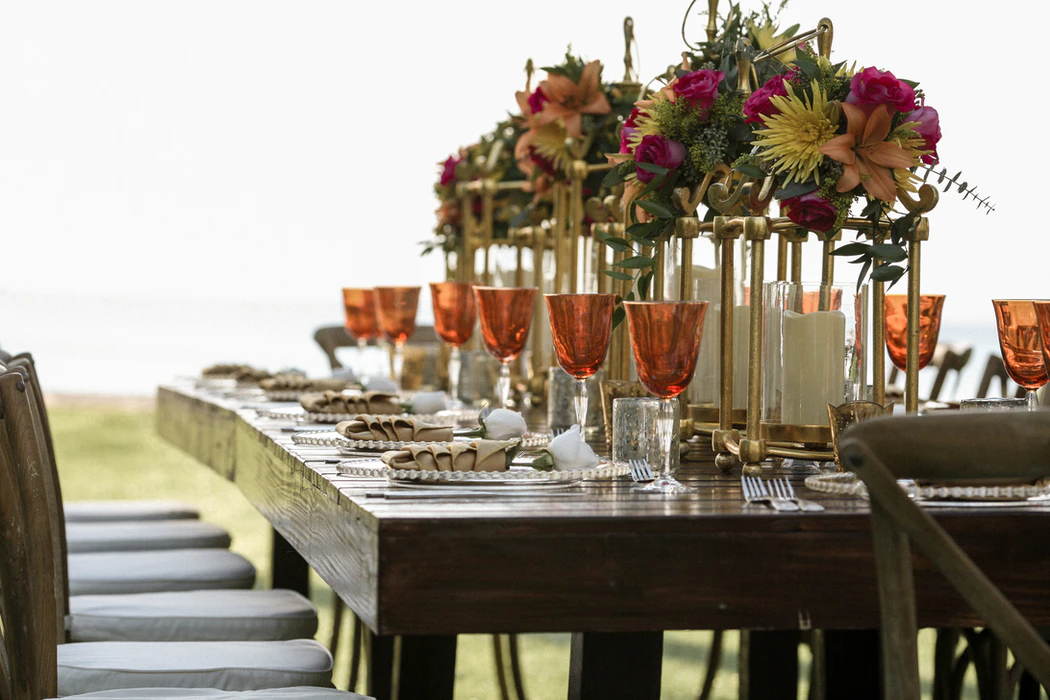 This is a photo of a fully set table: plates with elegantly folded napkins on top, wine glasses filled to the brim with a reddish/pinkish liquid, and a center piece running down the length of the table of brass-y metal bars and flowers.