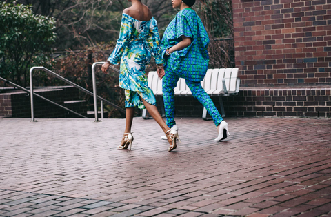 Two women walk on a brick path in brightly colored outfits, chic and stylish. Both are blue and green dominant