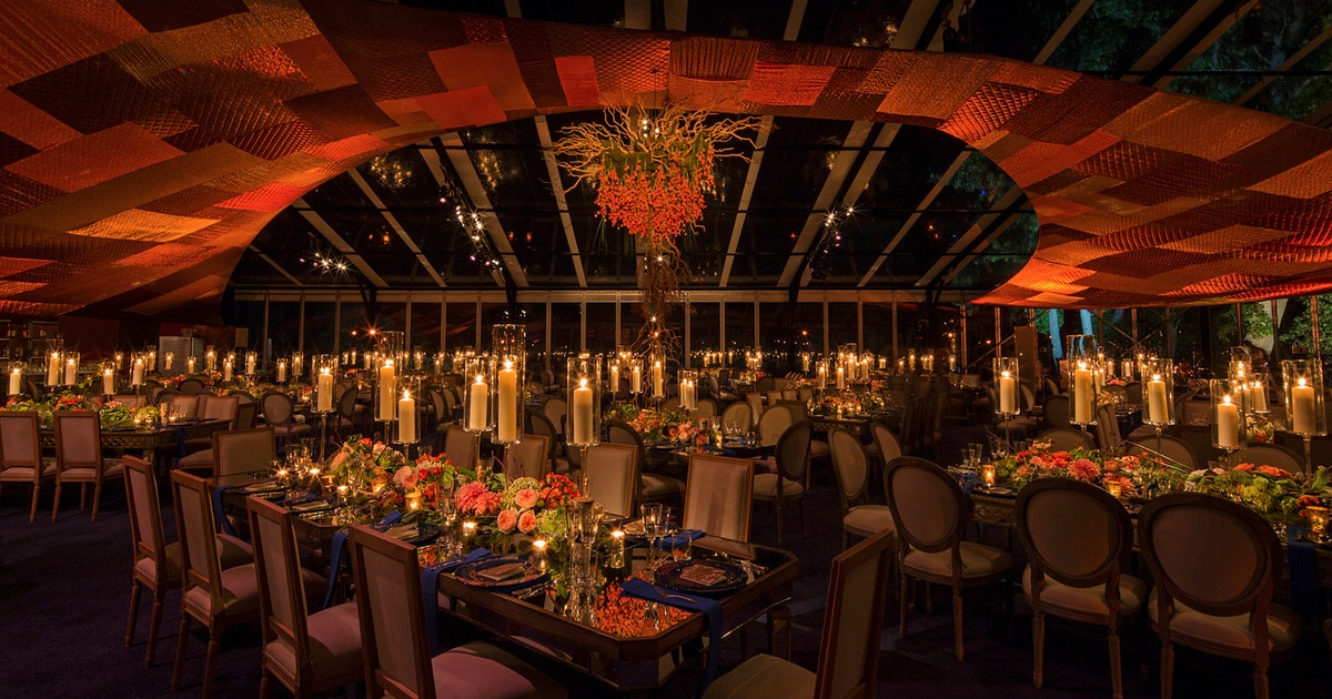 A large event space with numerous tables surrounded by chairs. Tables have full ornate place settings, covered in flower center pieces with lit candles. A red ceiling centers on an ornate flowery chandelier hanging from the ceiling.