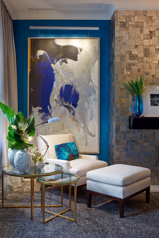 Corner of living room with nested side tables, an armchair with ottoman, and a large abstract blue painting on a blue wall