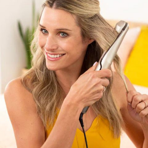 Une femme souriante utilisant un lisseur sur ses cheveux mi-longs et ondulés, habillée d'un top jaune.