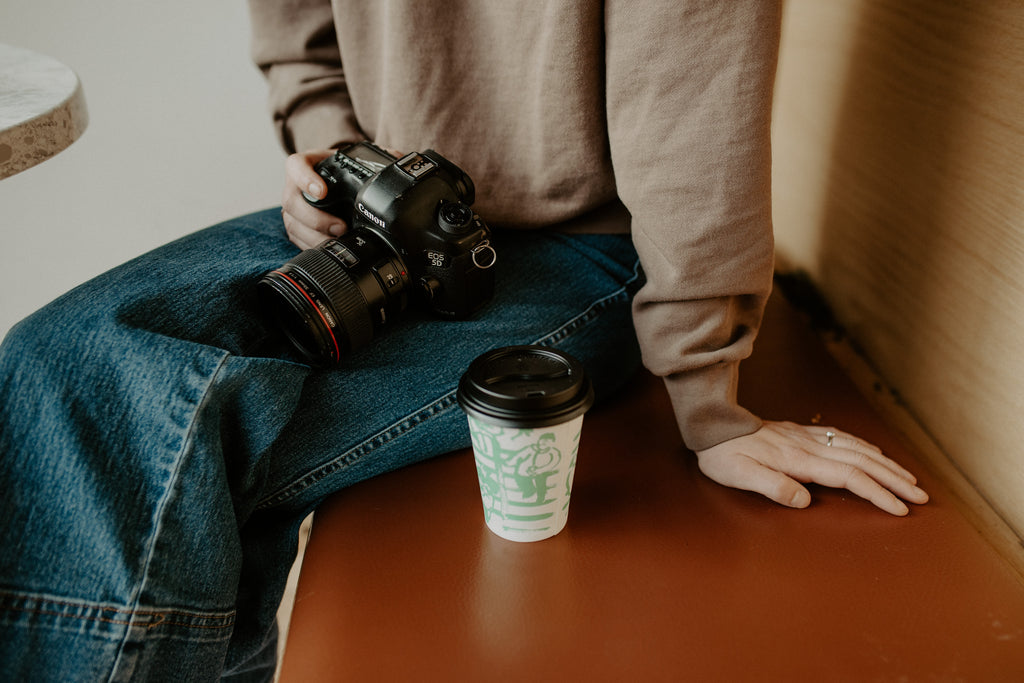 Coffee cup sits next to a girl holding a camera. Only lower torso is in view, sitting on leather bench