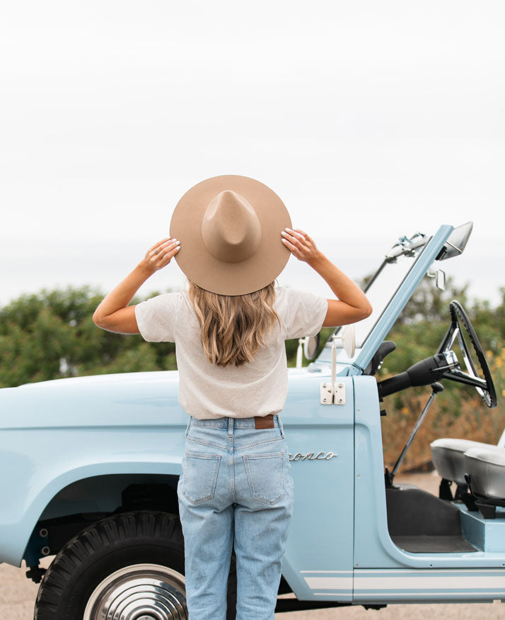 woman standing looking away from the camera, holding the brim of her hat and looking out towards the sea. in the background is a baby blue Ford Bronco.