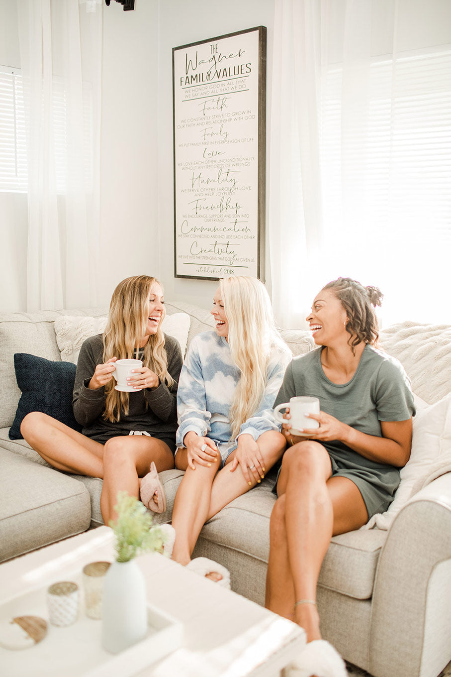 three female friends sitting on a couch and laughing while drinking out of mugs