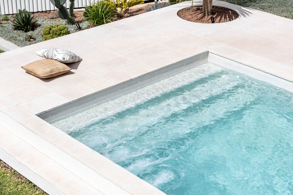 A swimming pool with blue tiles inside the pool and light beige marble tiles around the pool. It is a sunny day.