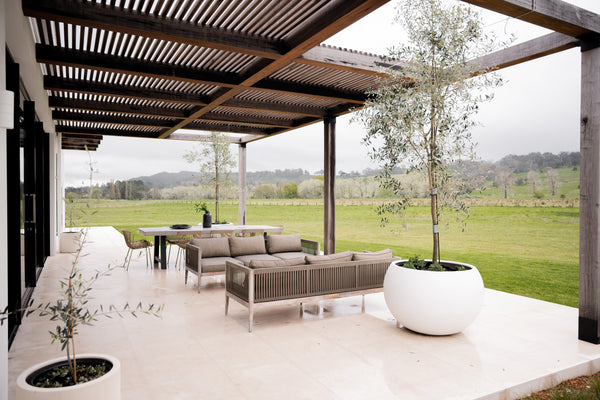 a modern style outdoor patio with beige marble on the floor, a pair of sofas. There is a white pot of flower next to the sofas. The patio area is covered with dark brown pergola.
