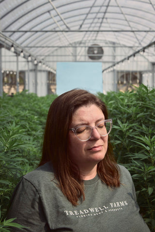 Jammie Treadwell inside a greenhouse wearing a Treadwell Farms shirt with her eyes closed standing in front of rows of hemp plants