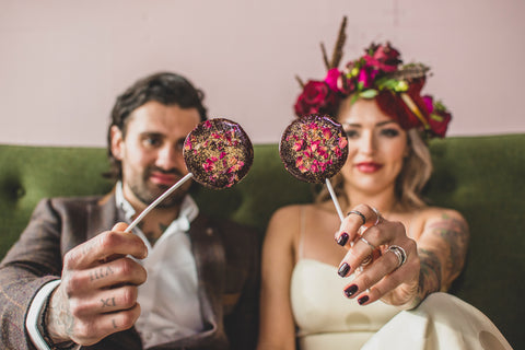 Alternative bride and groom holding black, rose and gold glitter wedding favour lollipops.
