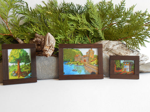 Three handmade miniature framed landscape drawing prints that are pictured around Thuya branches, driftwood and stones