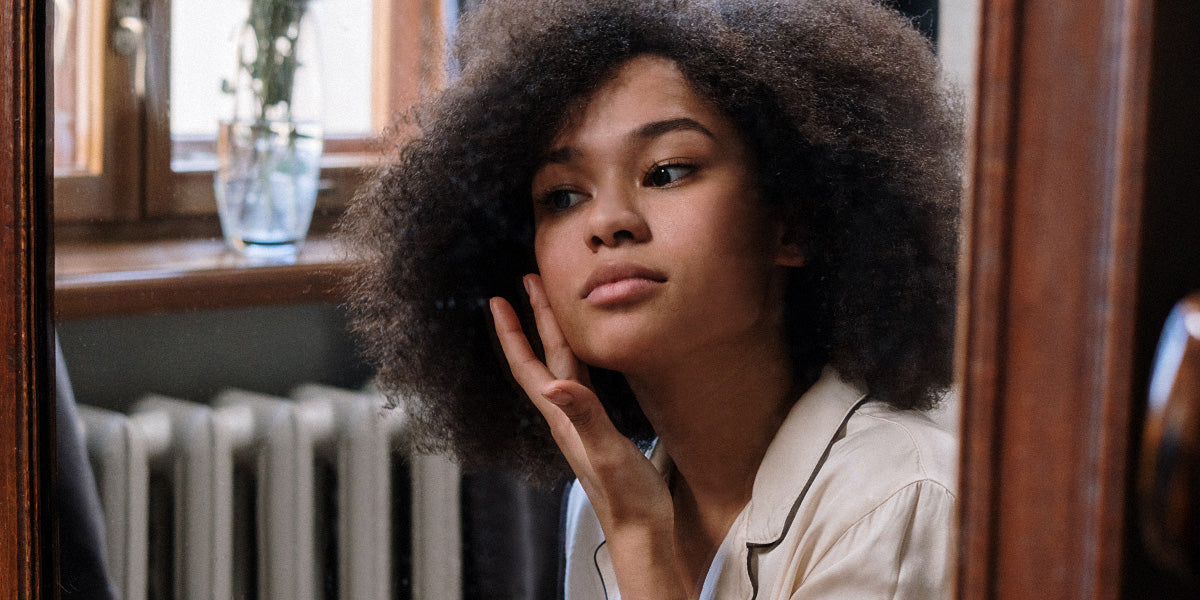 Frau mit dunkler Haut und Afro cremt ihr Gesicht sanft vor einem Spiegel ein