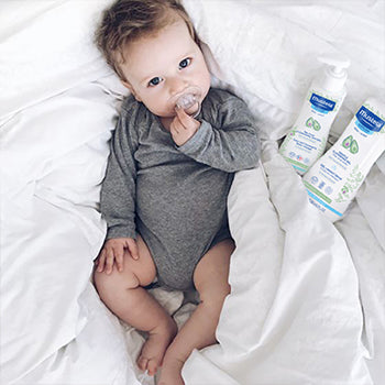 6 month old baby with a pacifier next to Mustela Products