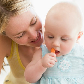 mother looking on as baby holds baby carrot at her mouth