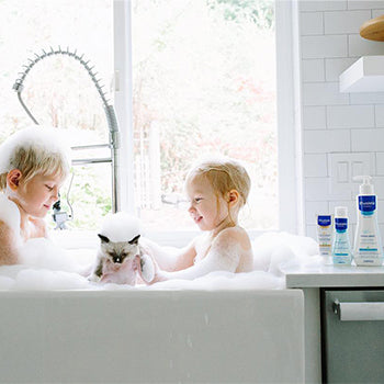 Dos niños pequeños bañándose en el fregadero de la cocina con un gatito y los productos de baño de Mustela