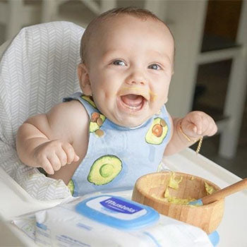 happy baby eating mashed avocado in a high chair with Mustela's cleansing wipes nearby