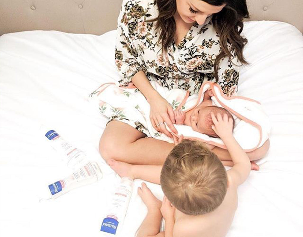 Mom holding newborn while sibling is on bed