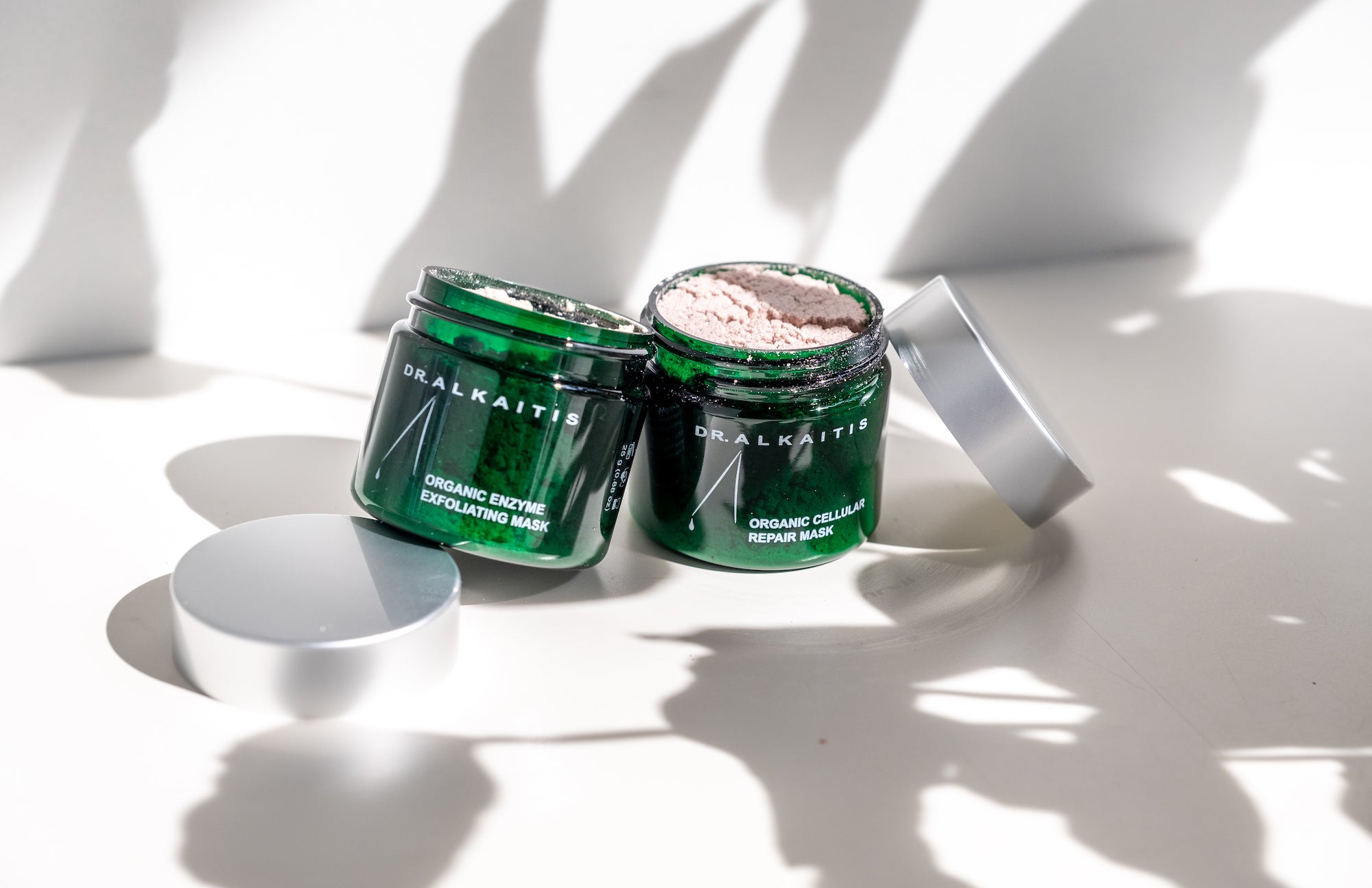 Dr Alkaitis Organics Cellular Repair Mask and Organic Enzyme Exfoliating mask on a white table with the shadows of leaves on them