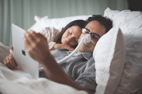 A man laying down and wearing a CPAP mask while looking at his tablet