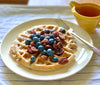 waffle on a plate with blueberries and pecans on top, with syrup on the side in a pitcher
