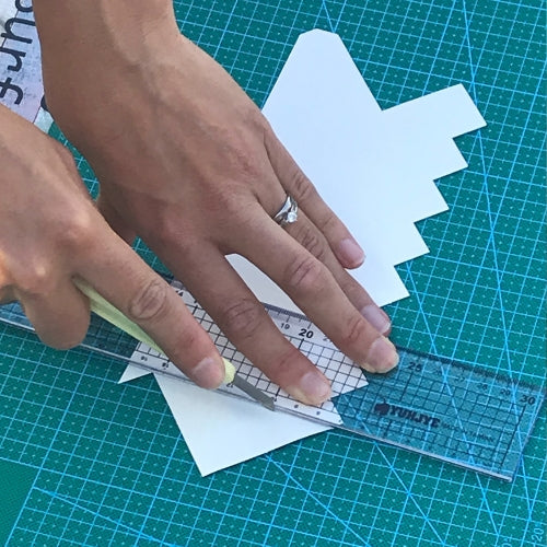 Hands cutting peel and stick tiles with craft knife along a metal edge ruler on a green craft mat