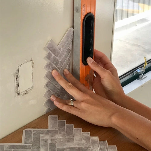 Hands checking angle of grey peel and stick chevron tiles with an orange level in caravan kitchen