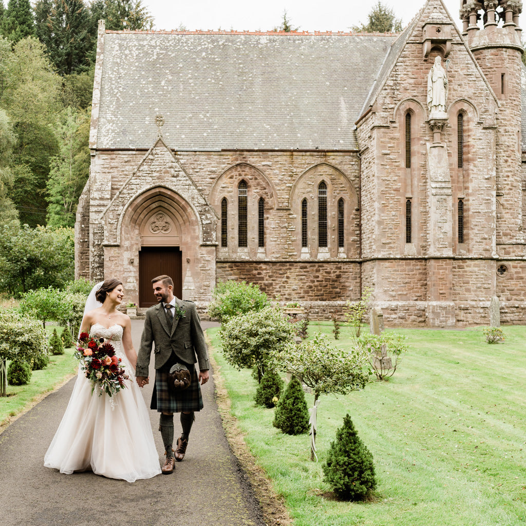 A just married couple walk in front of an old church with a woodland backdrop