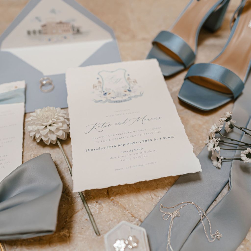 A dusty blue themed wedding stationery suite with an invitation with a custom wedding crest