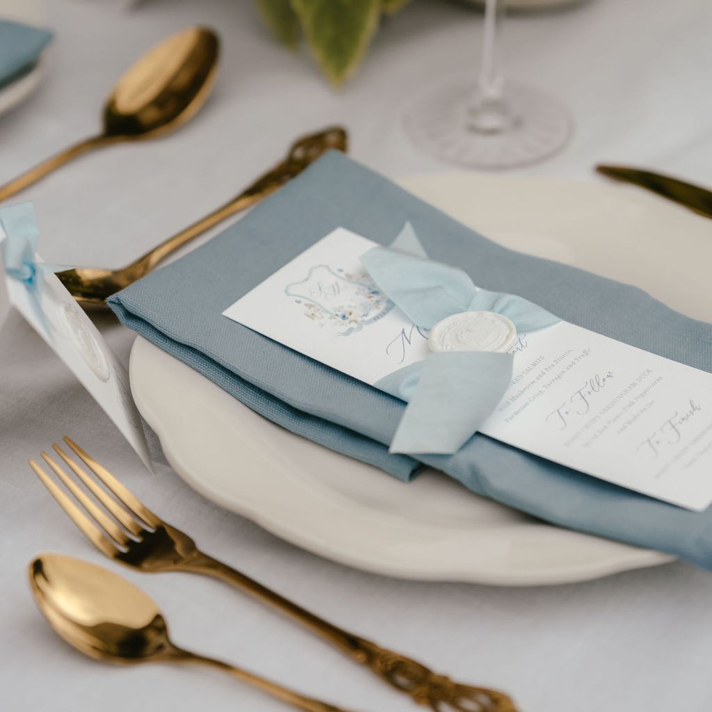 A photograph of a dusty blue themed wedding menu with a custom wedding crest