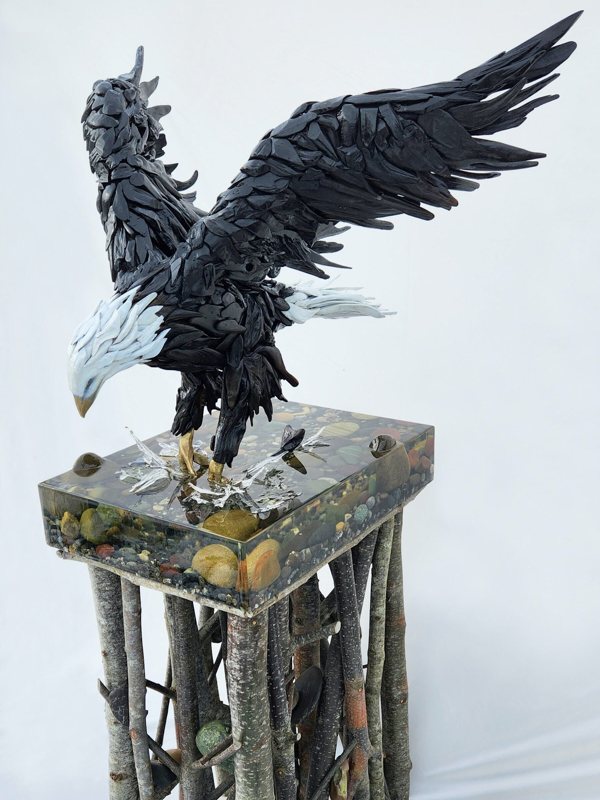 An intricate epoxy resin art piece featuring a bald eagle figurine sinking its talons into an epoxy layer representing a water stream to seize its prey.