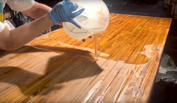 A large flood coat batch of epoxy resin being poured carefully onto the second half of a wooden bar top epoxy substrate.