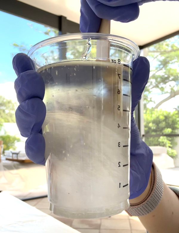 A small batch of UltraClear epoxy resin being mixed swiftly with a wooden stir stick in a small plastic measuring cup.