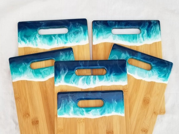Six different wooden cutting boards with an ocean resin art accent near each handhold.