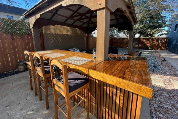 An outdoor wooden epoxy bar top with a wooden shelter above it and several wooden chairs surrounding it.